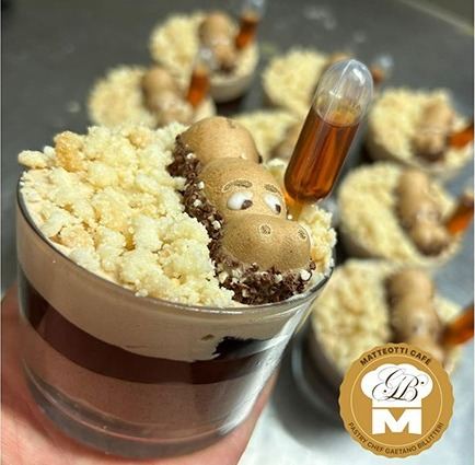Dessert al bicchiere con crema al cioccolato, crumble e decorazione, con pipetta di sciroppo; logo in basso a destra