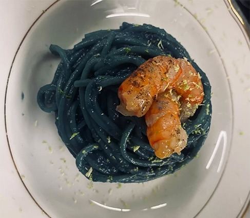 Spaghetti scuri con due code di scampo e scorza grattugiata, in un piatto fondo bianco con filo dorato.