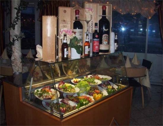 Banco espositivo refrigerato con piatti di antipasti e insalate, sopra bottiglie di vino e cassette di legno; sala sullo sfondo.